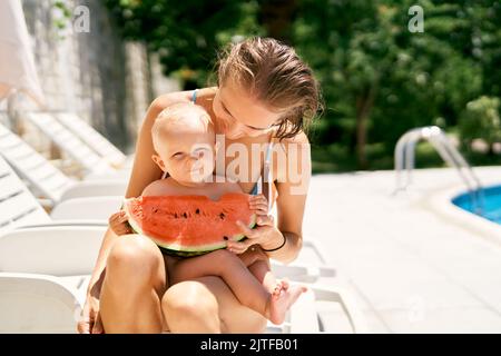Mama sitzt auf einer Sonnenliege und ein Baby hält eine Wassermelone Stockfoto