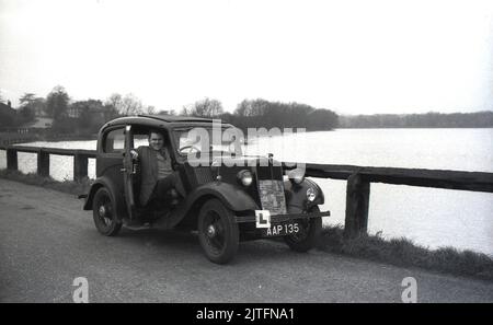 1950s, historisch, ein Mann, der mit geöffneter Tür in einem Vorkriegswagen, einem Morris Eight, auf einer schmalen Straße an einem großen See sitzt, England, Großbritannien. Der 1935 erstmals produzierte Eight, wie er allgemein bekannt war, war das beliebteste britische Auto der späten 1930s und wurde bis 1948 produziert. Durch seinen Erfolg wurde Morris Motors zum größten Motorenhersteller Großbritanniens. Stockfoto