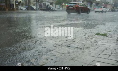 Regentropfen fallen auf die Straße, Autos fahren im Hintergrund. Starker sintflutartiger Regen in der Stadt. Stockfoto
