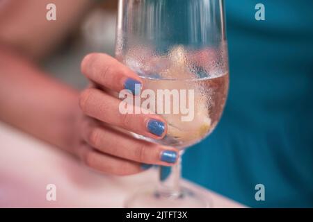 Die Hand einer jungen Frau hält ein Kristallglas mit Limonade und Zitrone in einer Bar mit Kleid und blau lackierten Nägeln Stockfoto
