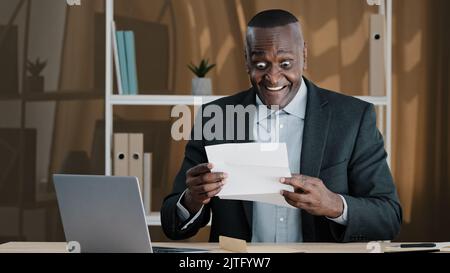 Alte ältere reifen biracial Chef Unternehmen CEO Arbeitnehmer erstaunt schockiert afrikanischen Mann Erfolg Geschäftsmann sitzen am Bürotisch offenen Umschlag lesen Brief Stockfoto