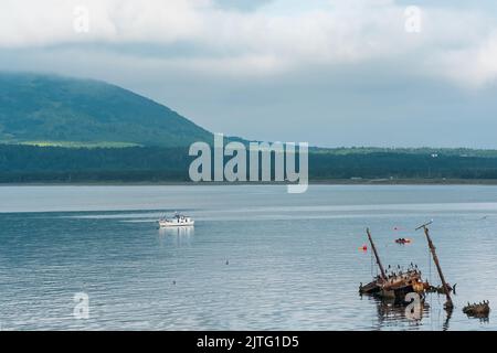 Wasserlandschaft der Meeresbucht mit einem Segelschiff und Schiffswrack und einem Berg in den Wolken im Hintergrund, Blick auf den Mendelejew Vulkan von der Seite Stockfoto