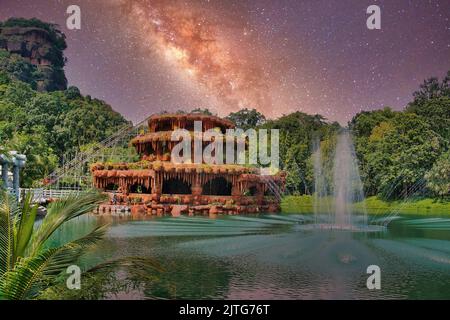 Fantasy-Landschaft mit einem surrealen, Tropfsteinhöhlenartigen Gebäude in einem See mit einem Brunnen, umgeben von Bäumen und einem hoch aufragenden Felsen, unter einem Sternenhimmel. Stockfoto