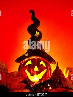 Ein glühender Jack o'Lantern mit einem langen schwarzen Hut, umgeben von Kürbissen und herbstgelben Blättern auf dunklem Grund. Abstraktes, minimalistisches Poster-Design f Stockfoto