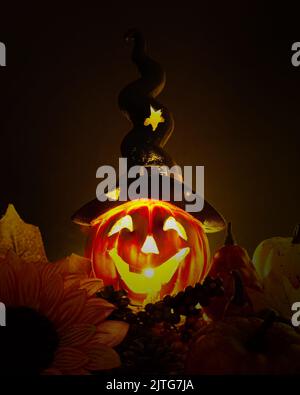 Ein glühender Jack o'Lantern mit einem langen schwarzen Hut, umgeben von Kürbissen und herbstgelben Blättern auf dunklem Grund. Abstraktes, minimalistisches Poster-Design f Stockfoto