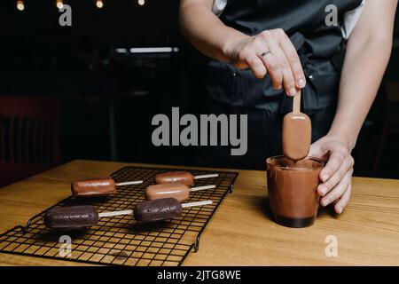 Eine Konditorin taucht einen Eiskuchen in geschmolzene Schokolade. Der Prozess der Herstellung von Cupcakes Stockfoto