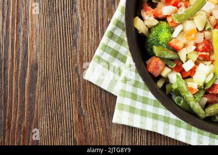 Eine Mischung aus verschiedenen gefrorenen Gemüse in einer Pfanne bereit zum Kochen auf braunem Holztisch. Draufsicht, flach liegend mit Platz für Text Stockfoto