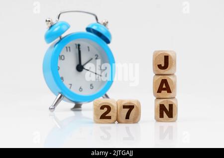 27.. Januar. Holzwürfelkalender für Januar 27, neben einem blauen Wecker. Objekte auf einer reflektierenden weißen Oberfläche Stockfoto