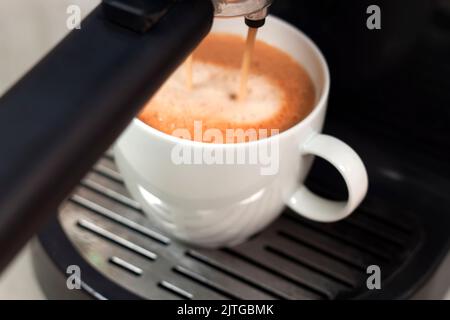 Glasbecher steht auf dem Gitter der schwarzen Kaffeemaschine und gießt Kaffee hinein. Prozess der Zubereitung von Kaffee. Stockfoto