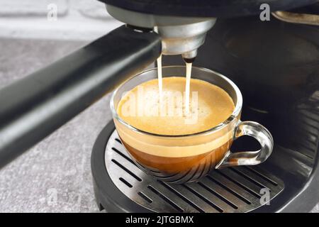 Glasbecher steht auf dem Gitter der schwarzen Kaffeemaschine und gießt Kaffee hinein. Prozess der Zubereitung von Kaffee. Stockfoto