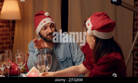 Glückliche Familienmitglieder tragen traditionelle Hüte und feiern den Winterurlaub zu Hause mit ihren Lieben. Festlicher Ehemann, der sich mit seiner schönen Frau unterhielt, während er beim Weihnachtsessen Champagner-Gläser anklickte. Stockfoto