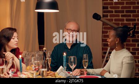 Fröhlicher älterer Mann, der sich mit einer jungen Frau unterhielt, während er am Weihnachtstisch mit Angehörigen zusammen war. Fröhlicher älterer Mensch, der mit seiner Tochter spricht, während er den Winterurlaub zu Hause gemeinsam feiert. Handgeführte Aufnahme Stockfoto