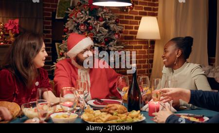 Festlicher Mann trägt Weihnachtsmann Kostüm auf Weihnachtsessen Party im Gespräch mit Familienmitgliedern. Junge Erwachsene Person, die als Weihnachtsmann getarnt ist und mit Menschen diskutiert, während sie den Winterurlaub feiert. Stockfoto