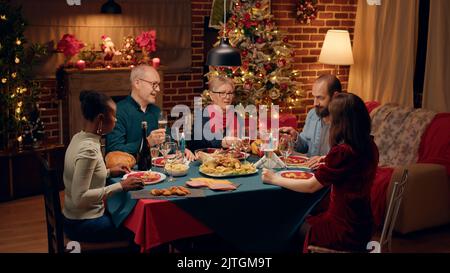 Lachend versammelten sich verschiedene Leute um den weihnachtlichen Esstisch und klimmten Weingläser. Fröhliche, herzlichst lächelnde Familienmitglieder feiern den traditionellen Winterurlaub zu Hause. Stockfoto