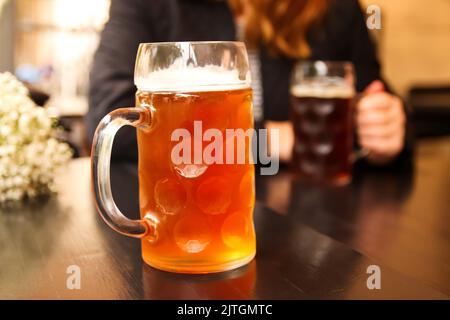 Defokussieren Sie den Becher mit leichtem Bier. Nahaufnahme von zwei Händen klirrenden Bier stein Gläser. Zwei Freunde trinken Bier im Pub. Nicht fokussiert. Stockfoto