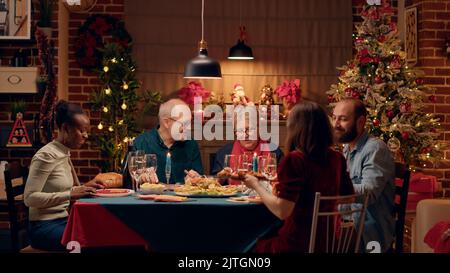 Positive multikulturelle Familienmitglieder feiern Weihnachten mit einem besonderen Abendessen zu Hause. Glückliche Menschen genießen traditionelle hausgemachte Speisen und haben eine gute Zeit zusammen. Stativaufnahme Stockfoto