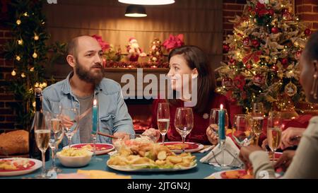 Festliches Paar feiert Weihnachten am Abendtisch mit engen Familienmitgliedern. Positive Menschen sitzen im Wohnzimmer, um authentische traditionelle Winterferien mit Angehörigen zu feiern. Stockfoto
