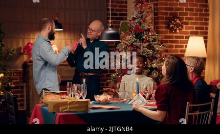 Fröhliche, vielfältige Familienmitglieder lachen herzlich zusammen, während sie das Weihnachtsessen zu Hause genießen. Lächelnde, herzhaft gelaunte Menschen genießen ein Winderfest, während sie sich unterhalten und sich gut fühlen. Stockfoto