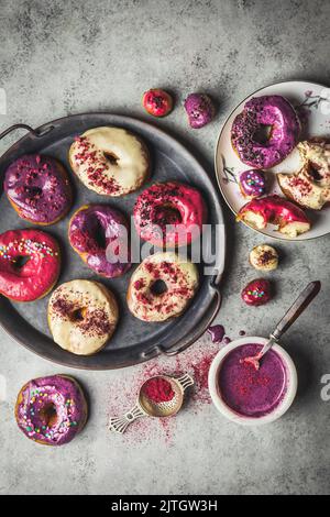 Donuts und Donut-Löcher mit beerfarbenen Glasuren und Streuseln auf Serviertablett mit Glasur in Schüssel Stockfoto