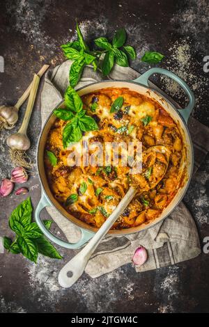 Gebackene Pasta mit Käsesauce und Würstchen im niederländischen Ofen, Basilikumgarnitur und Knoblauchköpfe auf der Theke Stockfoto