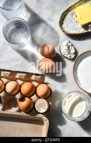Backzutaten im Sonnenlicht, darunter Eier, Mehl, Zucker und Butter Stockfoto