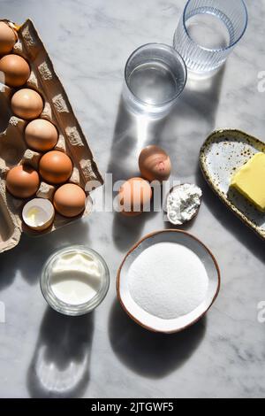 Backzutaten im Sonnenlicht, darunter Eier, Mehl, Zucker und Butter Stockfoto