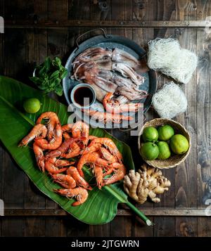 Zutaten für die thailändische Küche - Garnelen, Reisnudeln, Minze, grüne Zwiebeln, Tintenfisch, Limetten, Palmblatt-Komposition, Draufsicht Stockfoto