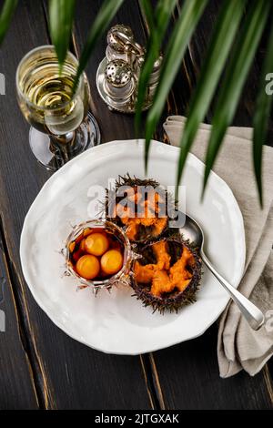 Frisch geöffnete Seeigel mit Wachtelei und Sojasauce auf weißem Teller auf schwarzem Holztisch Stockfoto