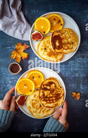 Mandelmilchpfannkuchen, serviert mit Orangenscheiben, Feigen und Schokoladenstückchen auf zwei weißen Tellern, Ahornsirup und Herbstblättern an der Seite. Frau hat sich einen Urlaub genommen Stockfoto