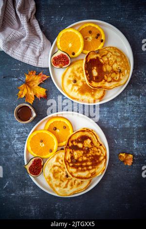 Mandelmilchpfannkuchen, serviert mit Orangenscheiben, Feigen und Schokoladenstückchen auf zwei weißen Tellern, Ahornsirup und Herbstblättern an der Seite. Stockfoto
