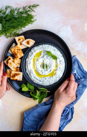 Frau mit türkischem Haydari-Meze-Dip, gekrönt mit frischen Minzblättern und Olivenöl, serviert auf schwarzen Tellern, begleitet von geschnittenem Pita-Brot. Stockfoto