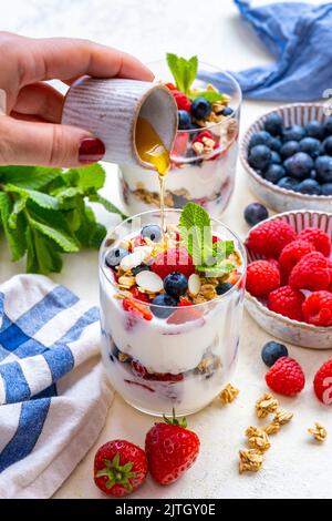 Frau gießt Ahornsirup über Joghurt-Parfait in einem Glas, Heidelbeeren, Himbeeren, Erdbeeren und Haferflocken in Keramikschüsseln auf der Seite. Stockfoto
