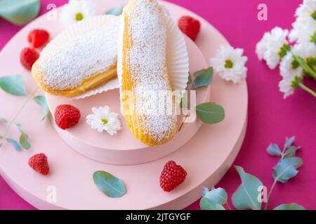 Eclairs Kuchen und Himbeeren auf einem rosa Podium auf einem fuchsia Hintergrund.Choux Gebäck. Sommerdesserts. Stockfoto