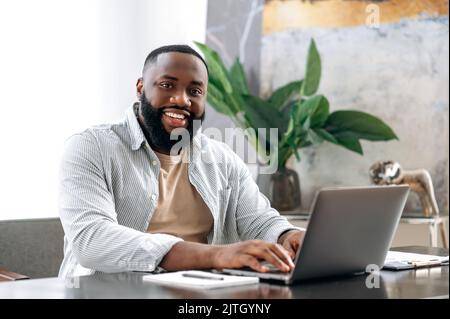 Porträt eines positiv selbstbewussten erfolgreichen afroamerikanischen jungen Mannes, Vertriebsleiter, ceo oder Freiberufler, sitzt an einem Schreibtisch mit Laptop im modernen Büro, blickt auf die Kamera und lächelt freundlich Stockfoto