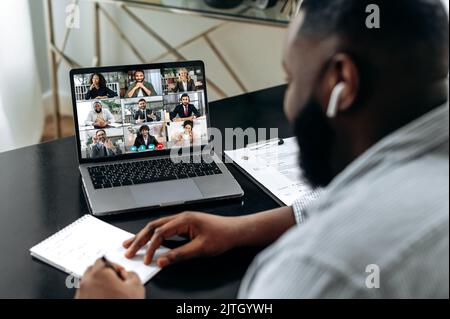 Ein freundlicher afroamerikanischer Mann, der sich an einen Schreibtisch setzt, ein Videotreffen mit Kollegen führt, ein finanzielles Brainstorming mit Geschäftspartnern durchführt, Notizen macht. Gruppenvideo-Treffen mit Menschen verschiedener Rassen Stockfoto