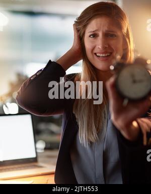 Im läuft die Zeit aus. Porträt einer verzweifelten jungen Geschäftsfrau, die im Büro steht und einen Wecker hochhält. Stockfoto