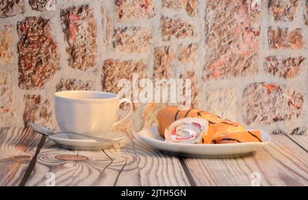 Ein Teller mit Biskuitrollen mit Creme und einer Tasse schwarzen Tee auf einem Holztisch. Nahaufnahme. Stockfoto