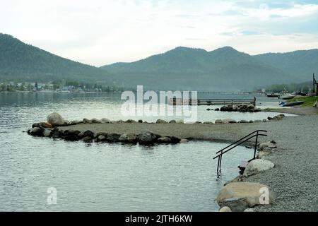 Metallgeländer von Treppen, die ins Wasser führen, und ein mit großen Steinen umzäunter Damm auf der Oberfläche eines großen Bergsees bei bewölktem Wetter. Stockfoto