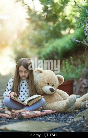Verloren im Wunder ihrer Lieblingsgeschichte. Ein kleines Mädchen liest ein Buch mit ihrem Teddybär neben sich. Stockfoto