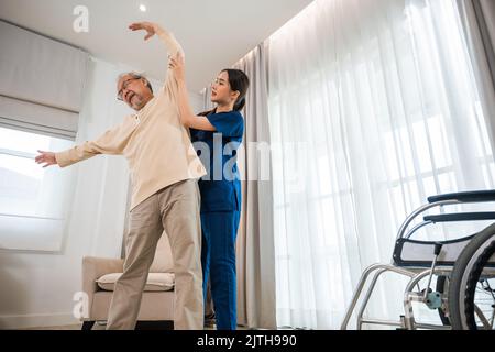 Der alte ältere Mann trainiert gerne bei einem Physiotherapeuten für ausgestreckte Arme zu Hause, der asiatische Physiotherapeut-Patient stillt ältere Menschen beim Trainieren Stockfoto