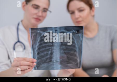 Der Arzt konsultiert eine kaukasische Frau und kommentiert die Röntgenaufnahme der Lunge. Stockfoto