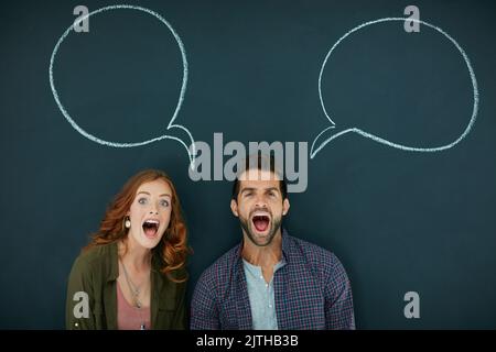 Wir haben viel zu berichten. Porträt eines jungen Paares, das vor einer Tafel steht, auf der Sprechblasen gezeichnet sind. Stockfoto