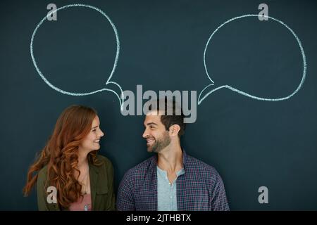 Erzählen Sie mir, was auf Ihrem Kopf ist. Ein junges Paar, das vor einer Tafel steht und auf der Sprechblasen gezeichnet sind. Stockfoto