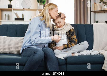 Reife blonde Frau, die nach der Messung die Temperatur ihres kranken Jungen auf dem Thermometer überprüft. Trauriges, krankes Kind, schaut mit seiner Mutter auf Thermometer. Stockfoto