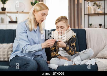 Reife blonde Frau, die nach der Messung die Temperatur ihres kranken Jungen auf dem Thermometer überprüft. Trauriges, krankes Kind, schaut mit seiner Mutter auf Thermometer. Stockfoto