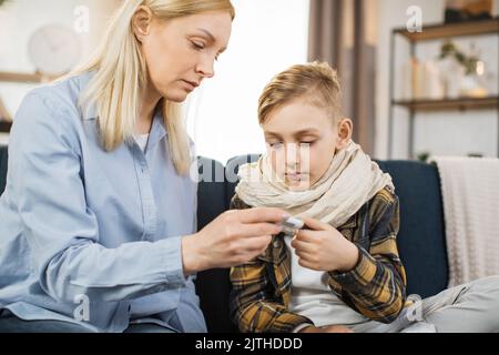 Reife blonde Frau, die nach der Messung die Temperatur ihres kranken Jungen auf dem Thermometer überprüft. Trauriges, krankes Kind, schaut mit seiner Mutter auf Thermometer. Stockfoto