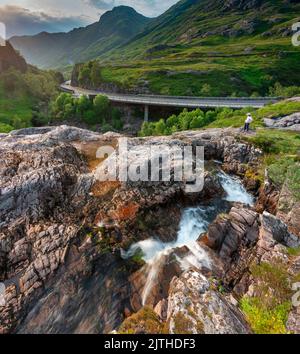 Glencoe, Schottland, UK-Juli 18. 2022: Eine Einzelfigur zeigt die Hauptstraße von Glencoe, während ein entferntes Auto aus dem verwinkelten Tal erscheint, bei Sonnenuntergang während der Sonne Stockfoto