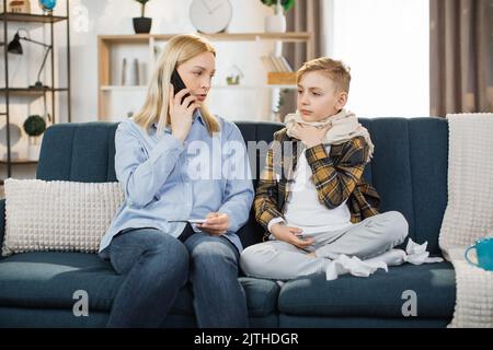 Familien-, Gesundheits- und Personenkonzept. Portrait einer blonden Mutter mit Thermometer und Smartphone, die zu Hause zum Arzt ruft. Kranker Sohn sitzt auf dem Sofa und hustet in der Nähe seiner Mutter. Stockfoto