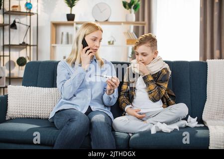 Familien-, Gesundheits- und Personenkonzept. Portrait einer blonden Mutter mit Thermometer und Smartphone, die zu Hause zum Arzt ruft. Kranker Sohn sitzt auf dem Sofa und hustet in der Nähe seiner Mutter. Stockfoto