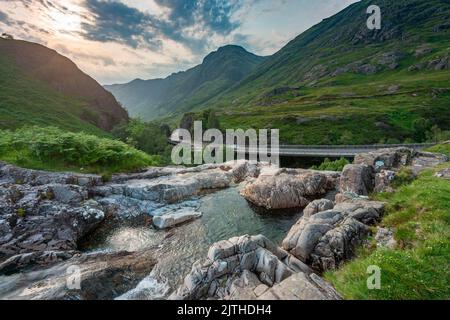 Die untergehende Sonne, über dem dramatischen Tal von Glen Coe, im Sommer, wo sich drei kleine Flüsse treffen, am Fuße der Three Sisters of Glen Coe, mit AW Stockfoto